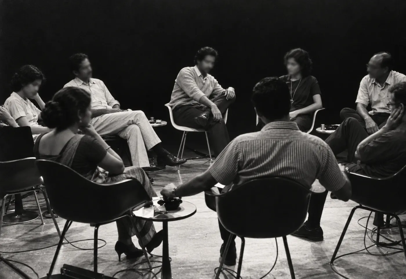 A circle of people in conversation on chairs around small tables, in black and white
